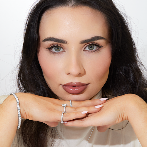 Woman with long dark hair and flawless makeup resting her chin on her hands, wearing diamond bracelets, a solitaire diamond ring, and a pavé band.