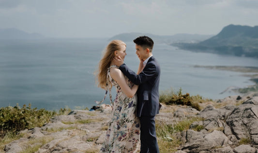 This image shows a couple standing close together on a rocky landscape, overlooking a scenic coastal view with mountains and the sea in the background. The woman wears a floral dress, and the man wears a formal suit. They're lovingly gazing at each other.