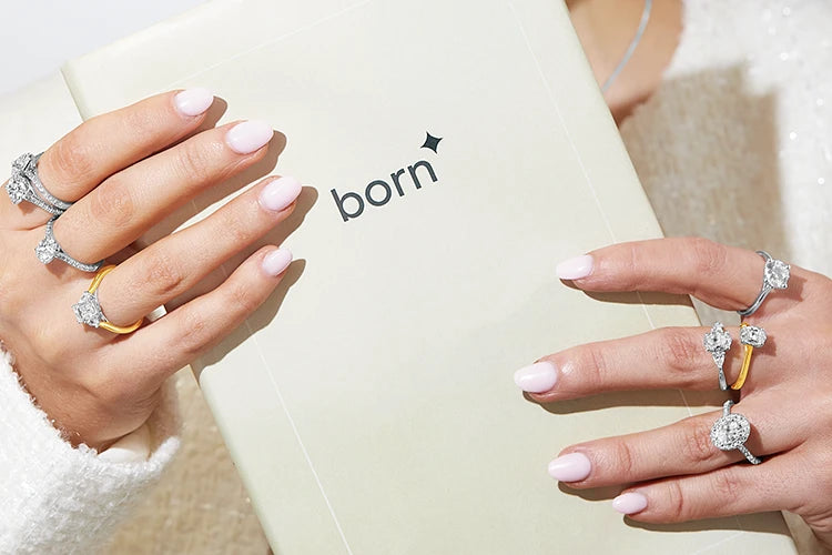Woman with pink manicured nails holding a "born" booklet, showcasing multiple diamond rings in white gold, yellow gold, and platinum on both hands. She wears soft makeup and a white textured top.