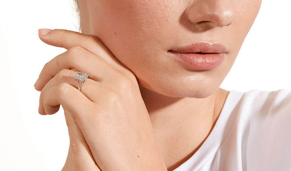 Close-up of a woman wearing a white top, gently touching her face with her hand, showcasing a sparkling radiant-cut diamond engagement ring.