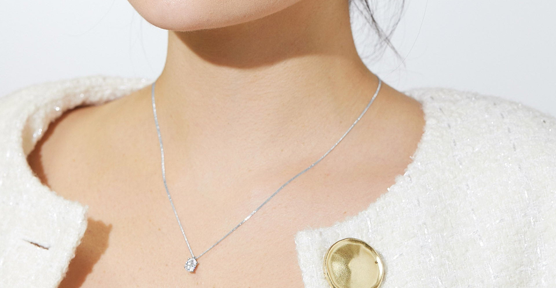 Woman wearing a cream textured jacket with a large gold button and a delicate pendant necklace featuring a single round diamond on a fine chain.