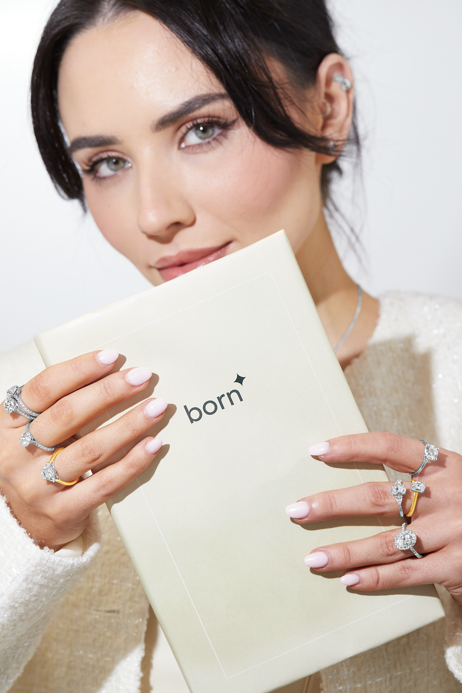 Woman with pink manicured nails holding a "born" booklet, showcasing multiple diamond rings in white gold, yellow gold, and platinum on both hands. She wears soft makeup and a white textured top.