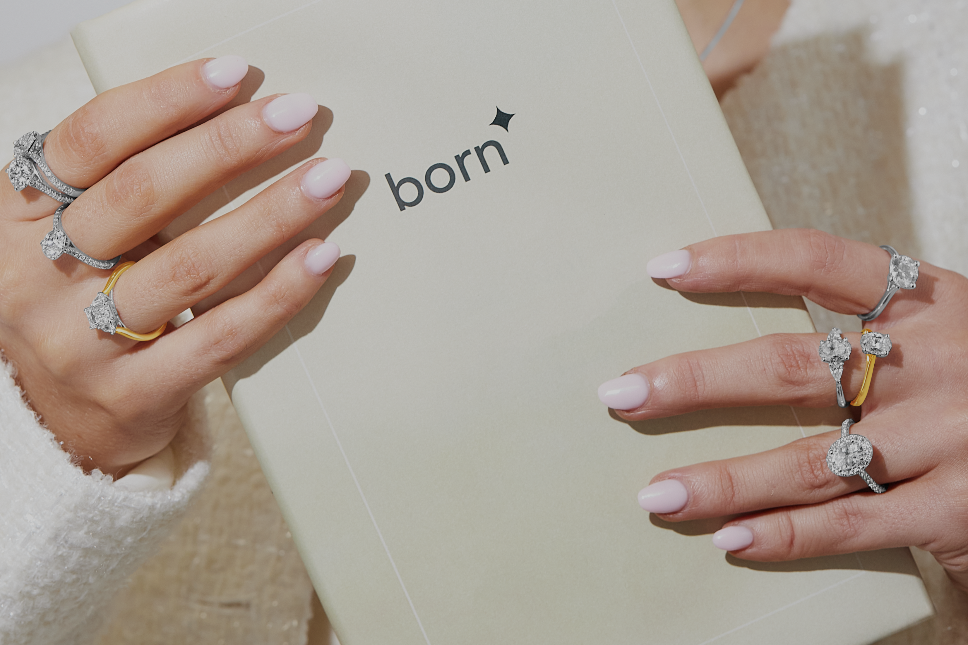 Woman with pink manicured nails holding a "born" booklet, showcasing multiple diamond rings in white gold, yellow gold, and platinum on both hands. She wears soft makeup and a white textured top.