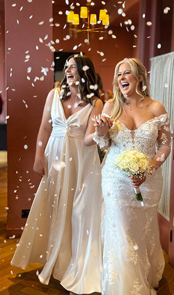 Two joyful brides walking hand in hand through falling flower petals. One wears an off-the-shoulder lace gown and holds a bouquet of white roses, while the other wears a satin gown with a deep V neckline.
