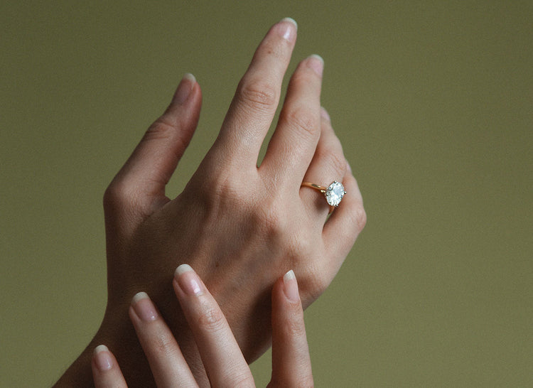 Woman with pink manicured nails holding a "born" booklet, showcasing multiple diamond rings in white gold, yellow gold, and platinum on both hands. She wears soft makeup and a white textured top.